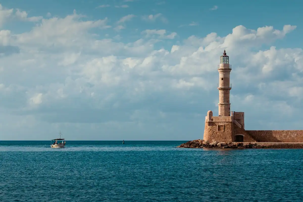 Far de piatră lângă un port, cu o barcă de pescari pe apă și nori deasupra