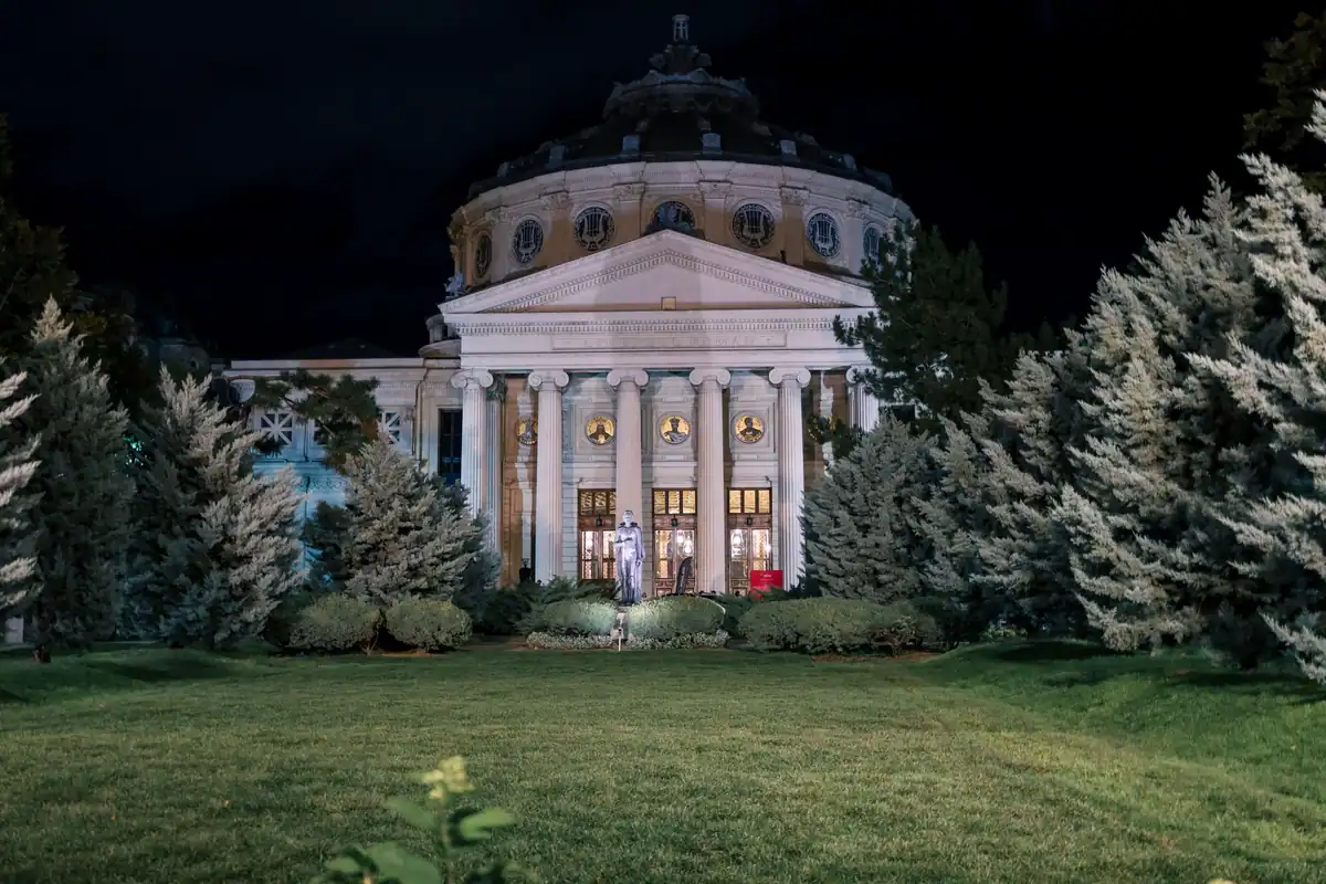 Ateneul Român iluminat noaptea, înconjurat de brazi, în centrul Bucureștiului