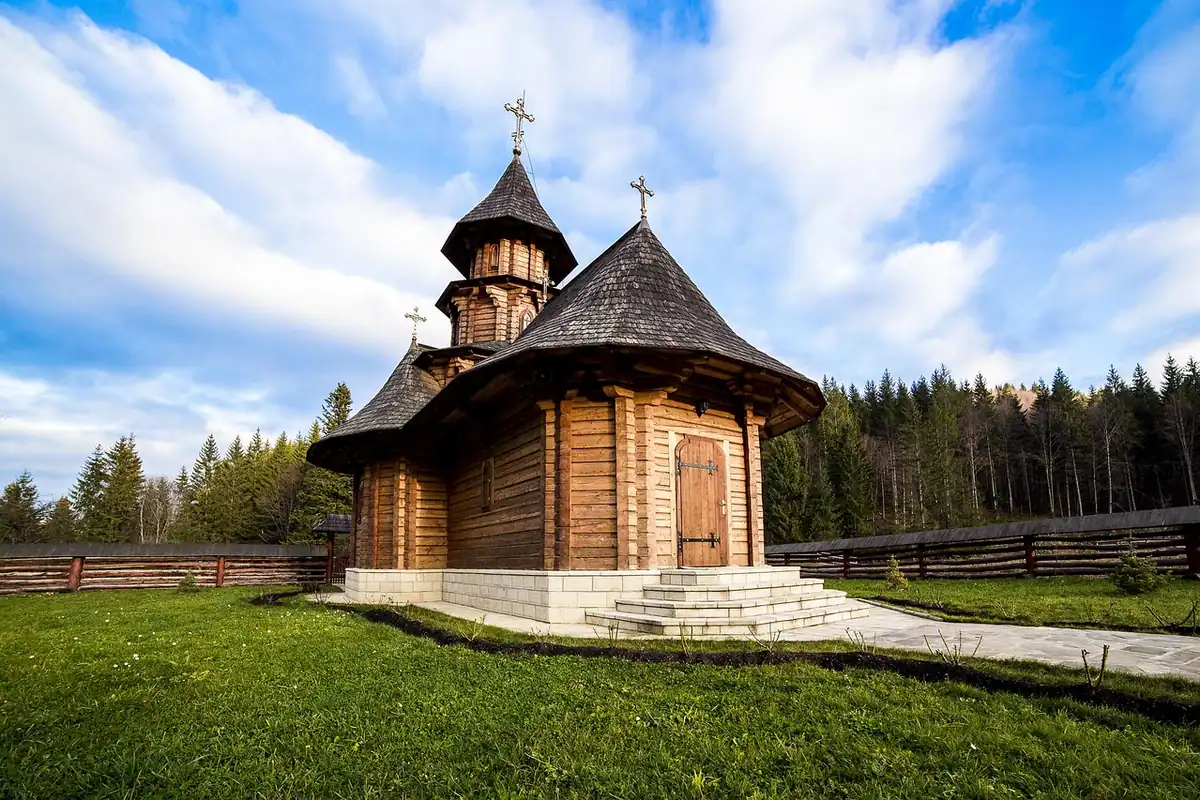 Biserică de lemn cu acoperiș în pante, pe un câmp verde, într-o mănăstire