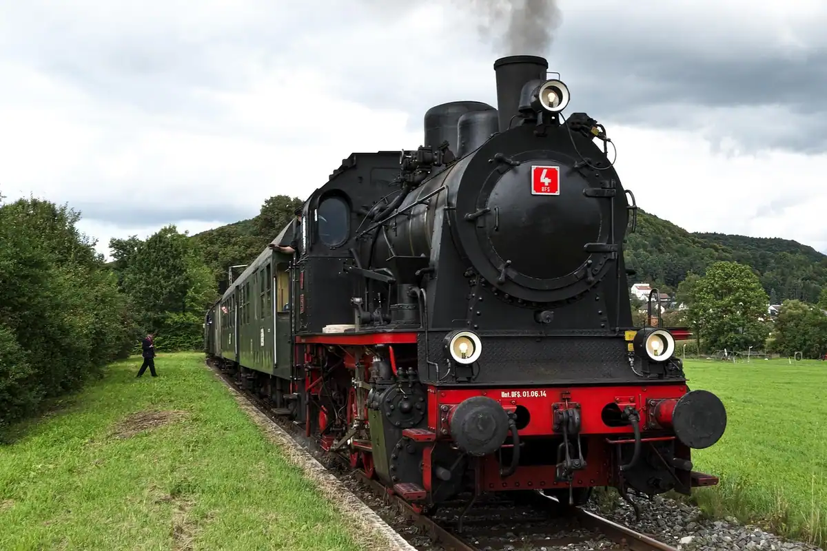 Locomotivă cu abur neagră, cu vagoane, pe o linie ferată prin câmp